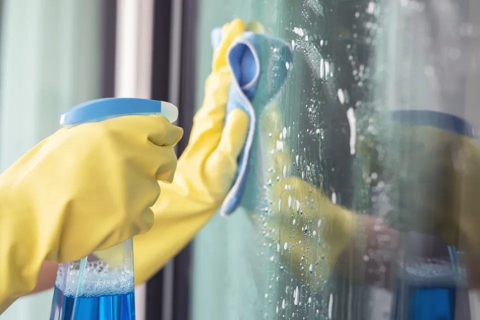 Gloved hands cleaning a glass window with a cloth and spray bottle
